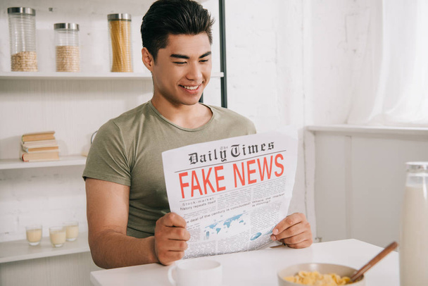 young asian man smiling while sitting at kitchen table and reading fake news newspaper - Valokuva, kuva