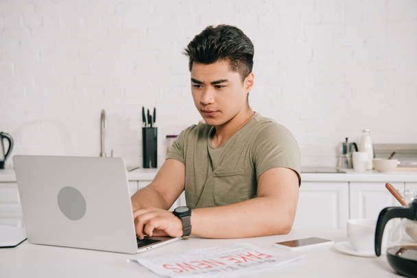 молодой, внимательный азиатский человек, использующий ноутбук, сидя за кухонным столом
 - Фото, изображение