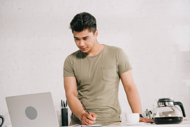 handsome asian man looking at laptop while standing near kitchen table - Фото, изображение