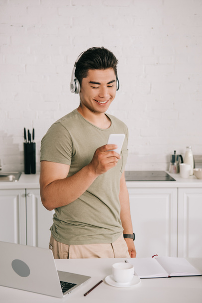 веселий азіатський чоловік слухає музику в навушниках, стоячи біля столу з ноутбуком і дивлячись на смартфон
 - Фото, зображення