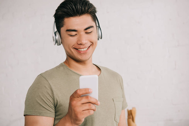 cheerful asian man listening music in headphones while looking at smartphone - Foto, imagen