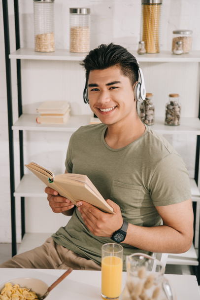 alegre asiático hombre en auriculares sonriendo a cámara mientras sentado en cocina mesa y celebración de libro
 - Foto, Imagen