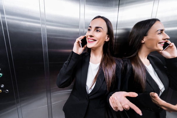 alegre mujer de negocios sonriendo y haciendo gestos mientras habla en el teléfono inteligente en el ascensor
 - Foto, Imagen