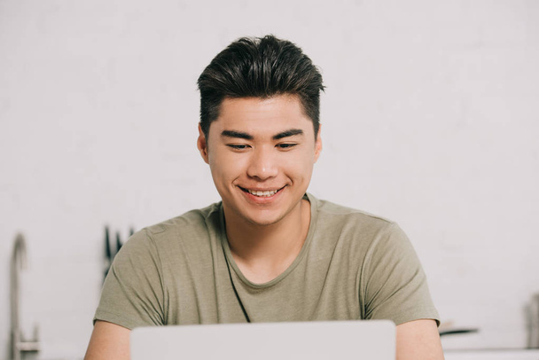 fröhliche asiatische Mann mit Laptop, während am Küchentisch sitzen - Foto, Bild