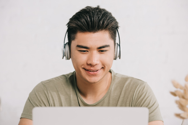fröhlicher asiatischer Mann benutzt Laptop und hört Musik über Kopfhörer - Foto, Bild