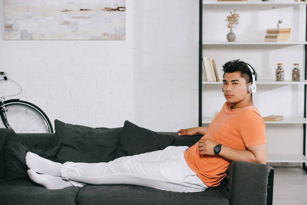 pensativo asiático homem em fones de ouvido olhando para câmera enquanto descansa no sofá em casa
 - Foto, Imagem