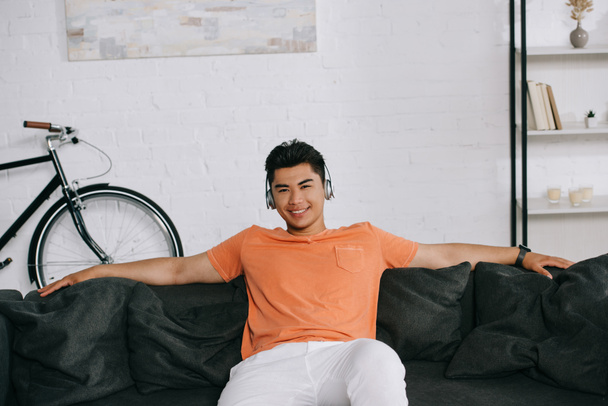 cheerful asian man smiling at camera while sitting on sofa and listening music in headphones - Photo, Image