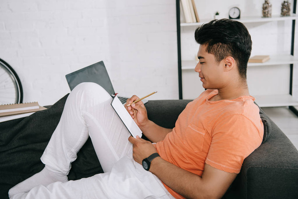young asian man writing in notebook while sitting on couch at home - Φωτογραφία, εικόνα