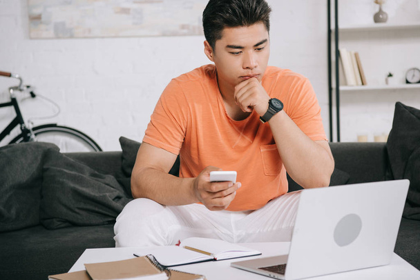 nachdenklicher asiatischer Mann schaut auf Laptop, während er auf Sofa sitzt und Smartphone in der Hand hält - Foto, Bild