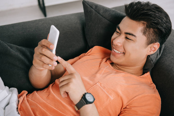 cheerful asian man using smartphone while relaxing on sofa at home - Valokuva, kuva