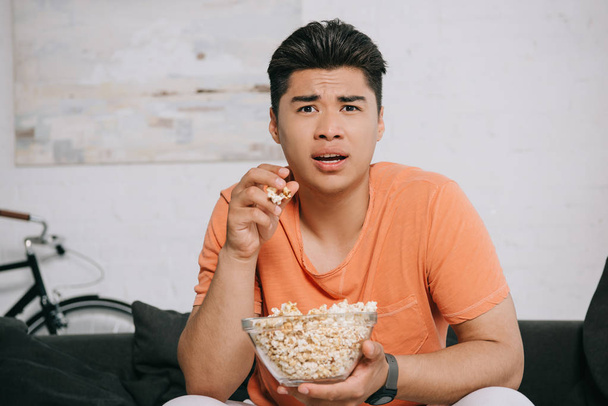 schockierter asiatischer Mann isst Popcorn und schaut fern, während er zu Hause auf dem Sofa sitzt - Foto, Bild