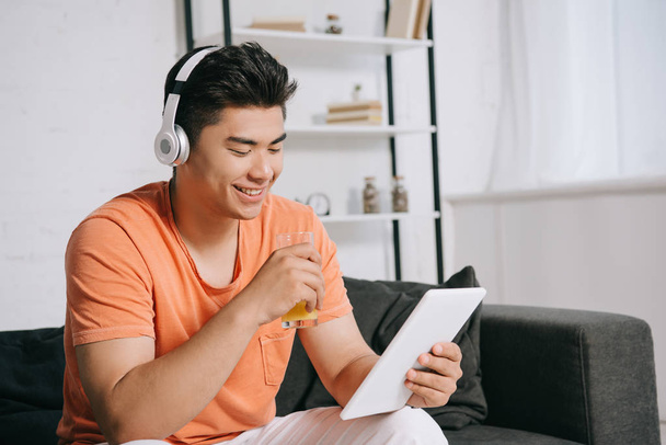 wesoły azjatycki człowiek picie soku pomarańczowego podczas korzystania z cyfrowego tabletu i słuchania muzyki w słuchawkach - Zdjęcie, obraz
