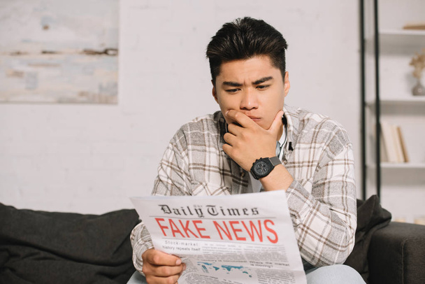 serious asian man reading fake news newspaper while sitting on sofa at home - Fotografie, Obrázek