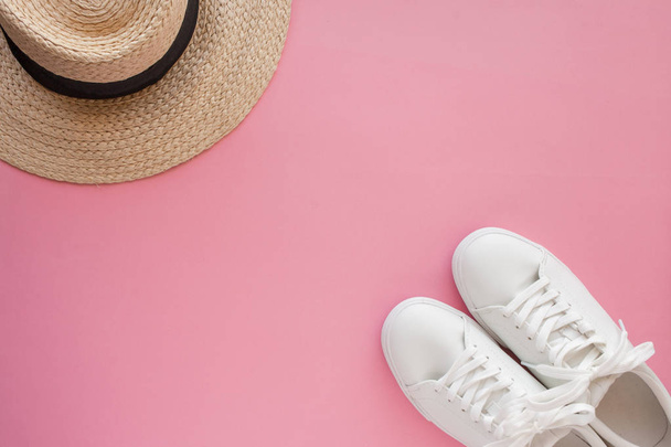 White sneakers lying on a pink background in front of a straw hat. - Foto, afbeelding