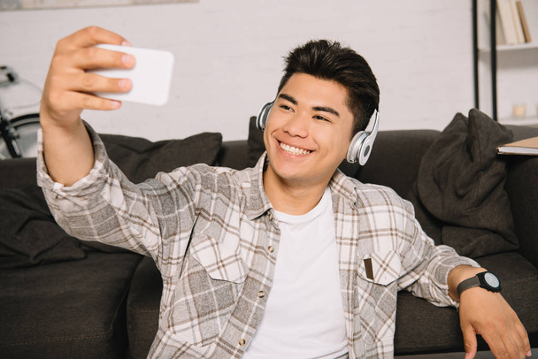 cheerful asian man in headphones taking selfie with smartphone while sitting on couch at home - Fotografie, Obrázek