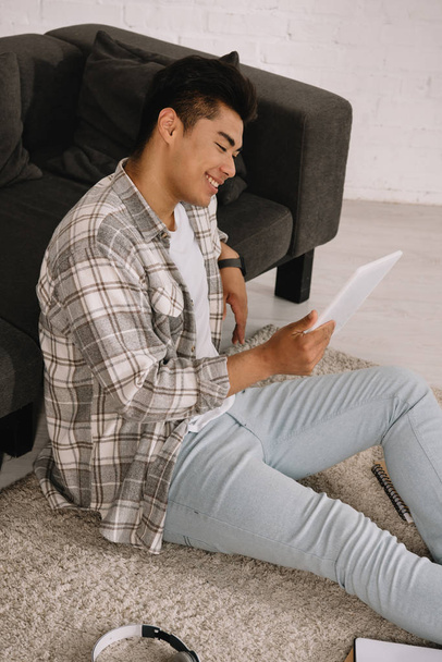 alegre asiático hombre usando digital tablet mientras sentado en piso cerca sofá en casa
 - Foto, Imagen