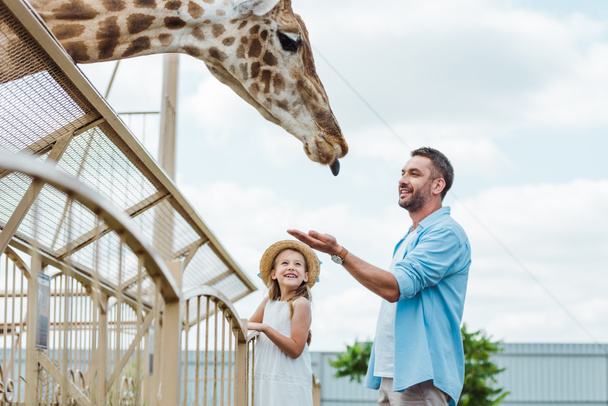 selective focus of cheerful man gesturing while looking at giraffe near kid in zoo  - Foto, afbeelding
