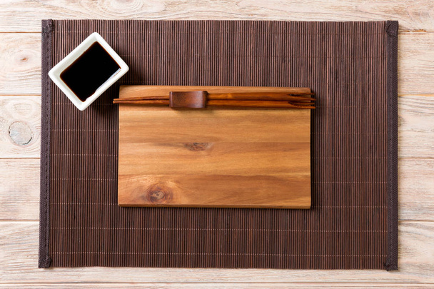 Пустая прямоугольная деревянная тарелка с палочками для суши и соевым соусом на деревянном фоне. Вид сверху с пространством для копирования
 - Фото, изображение