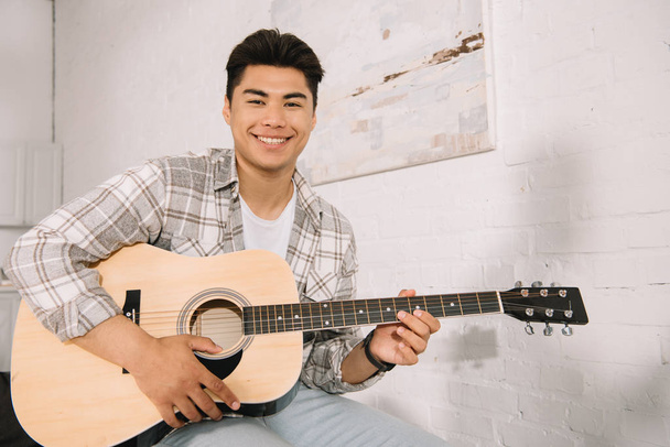 happy asian man smiling at camera while playing acoustic guitar at home - Foto, Imagem