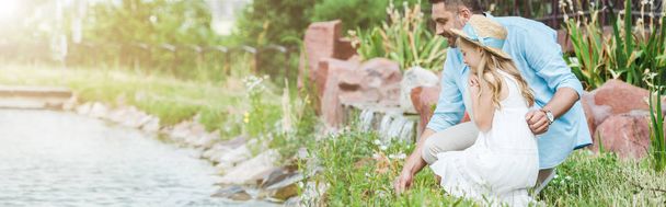 panoramic shot of cute kid in dress and straw hat sitting near happy father and lake  - 写真・画像