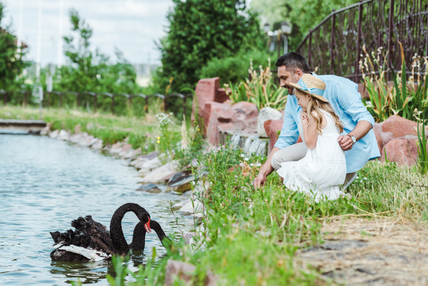 selective focus of cute kid in dress and straw hat sitting near happy father and lake with black swans  - Photo, Image