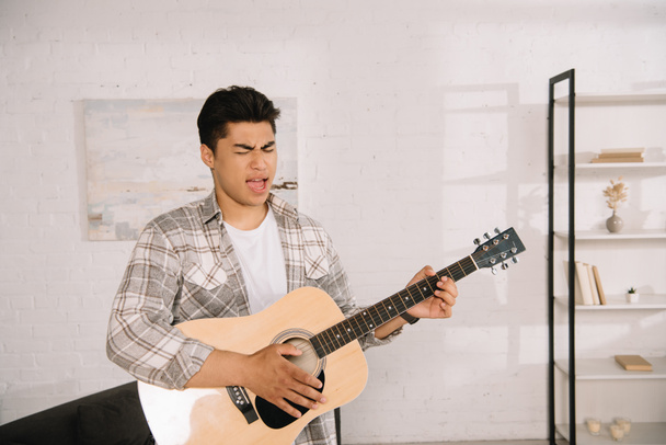 pohledný asijský muž hrající na akustickou kytaru a zpěv doma - Fotografie, Obrázek