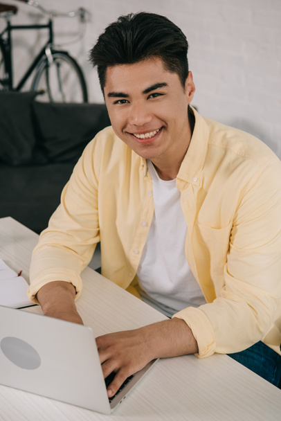 cheerful, young asian man smiling at camera while using laptop at home - Photo, Image