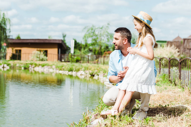 cute kid in dress and straw hat standing near happy father and lake  - Foto, Imagem