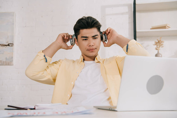 nachdenklicher asiatischer Mann mit Kopfhörern sitzt am Schreibtisch und schaut auf Laptop - Foto, Bild