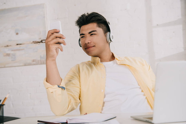 jonge, lachende Aziatische man in koptelefoon nemen selfie met smartphone  - Foto, afbeelding