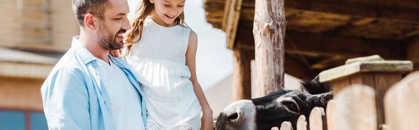 動物園の木製のフェンスの近くに腕かわいい娘を抱いて幸せな父親のパノラマショット  - 写真・画像