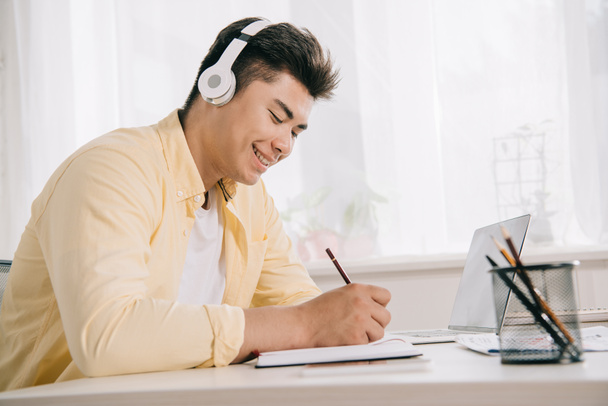 χαρούμενο ασιατικό άντρα στα ακουστικά γράφοντας στο σημειωματάριο ενώ κάθεται στο γραφείο - Φωτογραφία, εικόνα