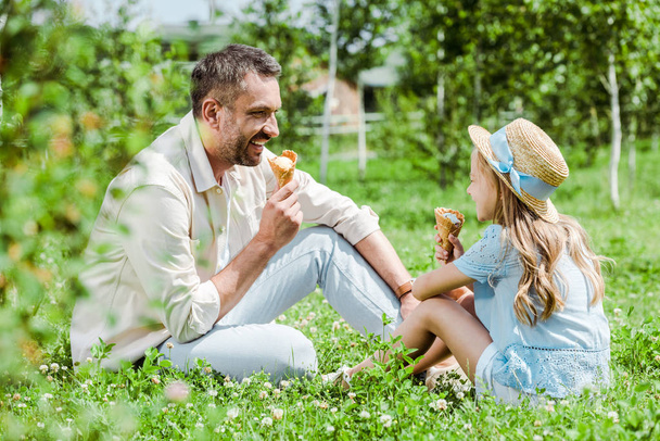 selective focus of cheerful father looking at kid in straw hat with ice cream cone sitting on grass  - 写真・画像