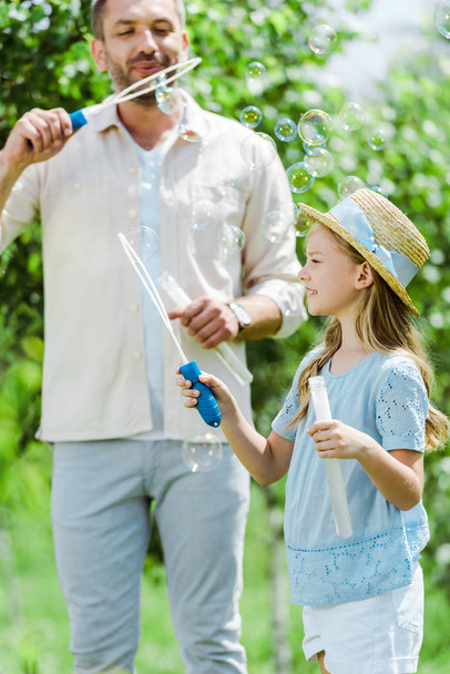 Selektiver Fokus der niedlichen Tochter im Strohhut mit Blasenstab in der Nähe des Vaters, der Seifenblasen pustet  - Foto, Bild