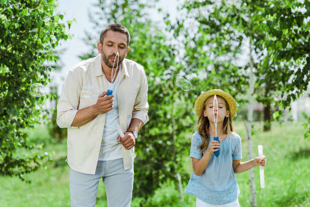 handsome man and daughter in straw hat blowing soap bubbles near trees - Fotografie, Obrázek