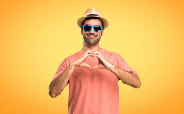 Homme avec un chapeau et des lunettes de soleil sur ses vacances d'été faisant symbole de coeur par les mains. Etre amoureux sur fond orange
 - Photo, image