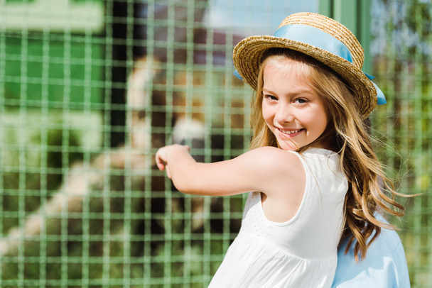 hasır şapkalı mutlu çocuk kameraya bakıyor ve hayvanat bahçesinde kafesin yakınında gesturing  - Fotoğraf, Görsel