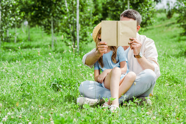 πατέρας και κόρη καλύπτοντας πρόσωπα κρατώντας το βιβλίο και καθισμένος στο γρασίδι  - Φωτογραφία, εικόνα