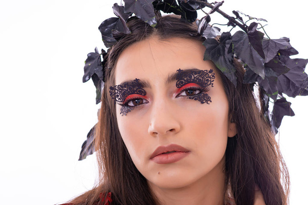 A vonzó fiatal női modell portréja. A lány pózol dekorációk a fejét egy stúdióban, fehér háttérrel. Fekete és piros levelek, mint a dekoráció. Fantasztikus make-up a szemét. - Fotó, kép