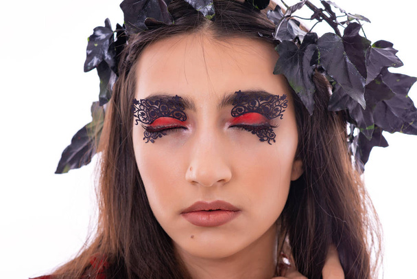 Portret atrakcyjne młoda kobieta model. Dziewczyna pozuje z dekoracjami na głowie w studiu z białym tłem. Czarne i czerwone liście jak dekoracja. Fantastyczne makijaż na oczach. - Zdjęcie, obraz