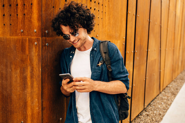 Close up portrait of handsome man standing outdoors, typing messages on mobile phone. Young male with curly hair wears sunglasses resting outside in the city browsing on his cell phone on modern wall - Фото, зображення