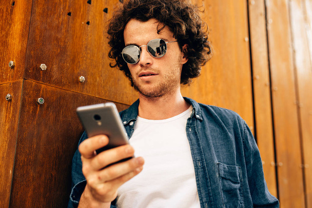 Κλείστε το πορτραίτο του όμορφου άντρα που στέκεται σε εξωτερικούς χώρους, πληκτρολογώντας μηνύματα στο κινητό τηλέφωνο. Νεαρό αρσενικό με σγουρά μαλλιά φοράει γυαλιά ηλίου που ξεκουράζονται έξω στην πόλη περιήγηση στο κινητό του στο σύγχρονο τοίχο - Φωτογραφία, εικόνα