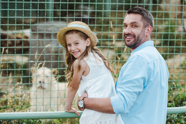 feliz papá y la hija mirando a la cámara cerca de la jaula con animales salvajes en el zoológico
  - Foto, Imagen