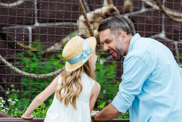 Kind mit Strohhut schaut Vater an und zeigt Zunge in Käfig mit Wildtier im Zoo  - Foto, Bild