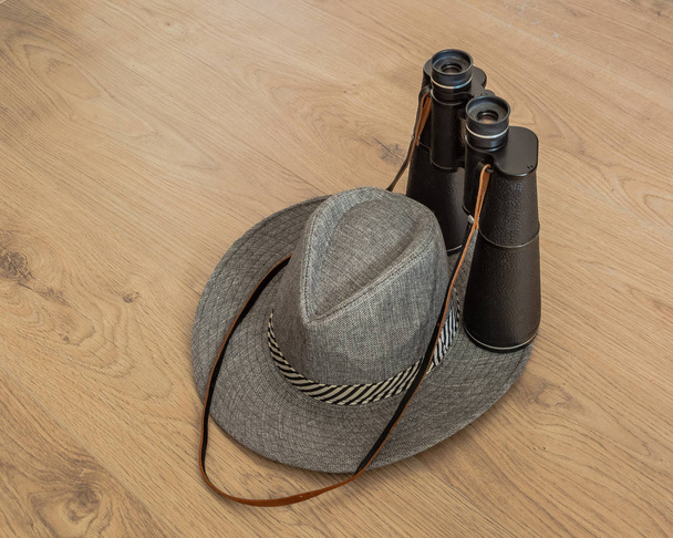 Wide-brimmed gray hat and binoculars, as symbols of travel. Wooden yellowish background. Preparation for the designer. Copy space. - Foto, afbeelding