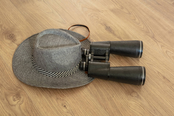 Wide-brimmed gray hat and binoculars, as symbols of travel. Wooden yellowish background. Preparation for the designer. Copy space. - Foto, Bild