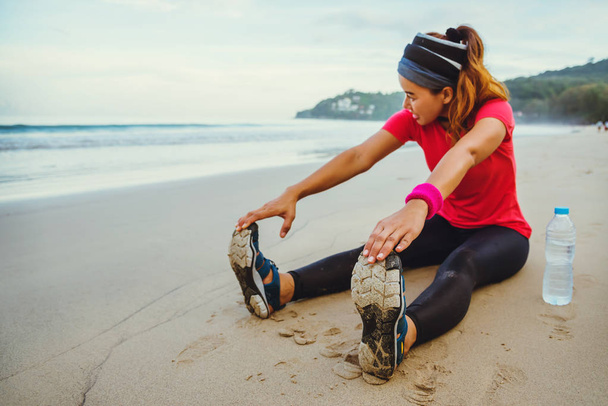 Asian Women jogging trening na plaży. Usiądź na plaży fitness relaks z rozciągliwy nogi i ramię Stretch. Jogging na plaży. Podróże Tajlandia, ćwiczenia. - Zdjęcie, obraz