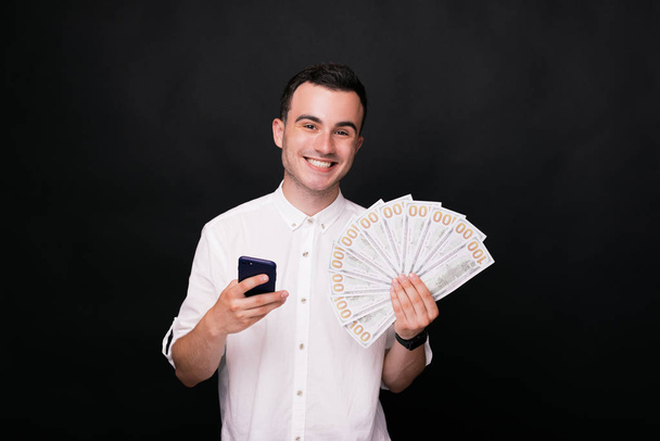 I won! Young smiling man at the camera, holding his phone is the other hand on black background in white shirt. - Φωτογραφία, εικόνα