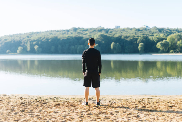 Young man standing with his back at the camera  at the shore of the lake watching the forest bathed in sunlight in the morning. - Fotografie, Obrázek