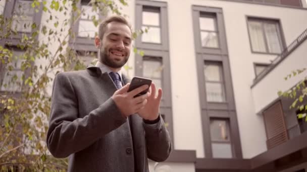 Happy handsome business man in coat using smartphone while standing outdoors in the city - Séquence, vidéo